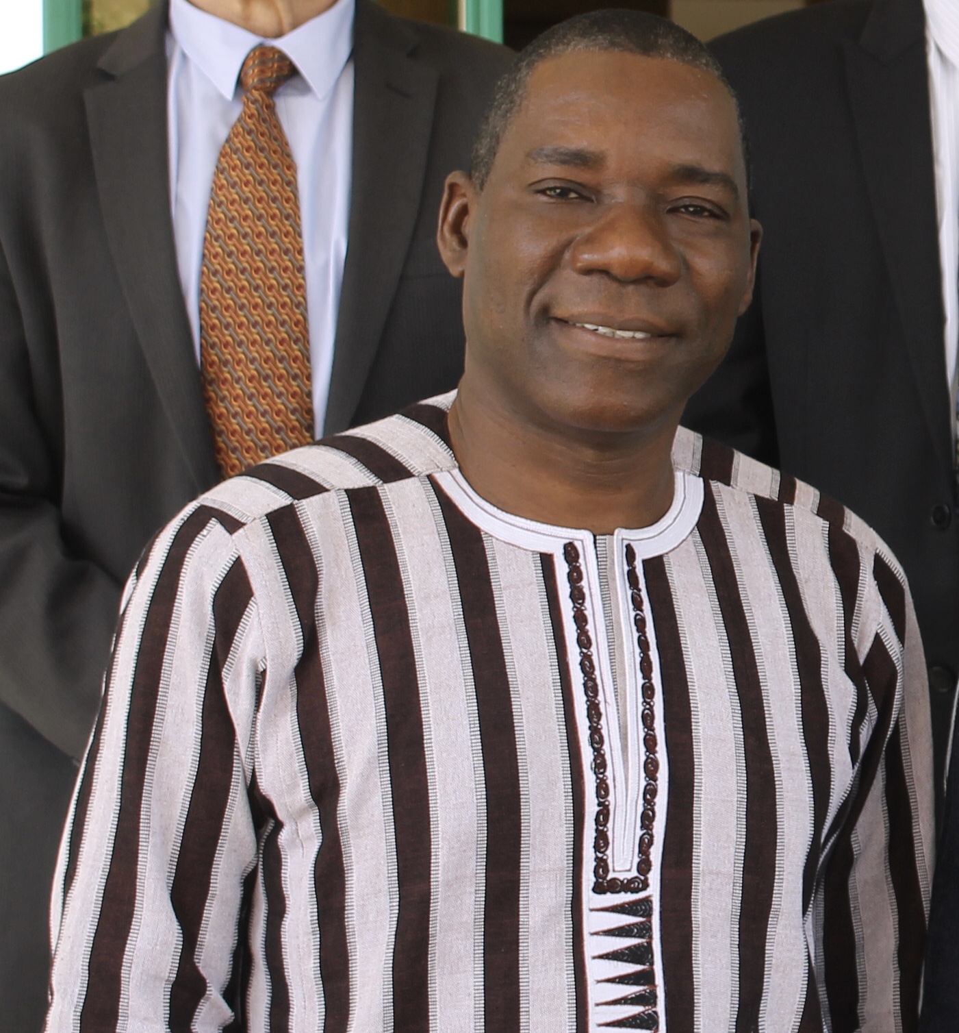 Homme souriant en tenue rayée traditionnelle debout devant deux hommes en costume cravate