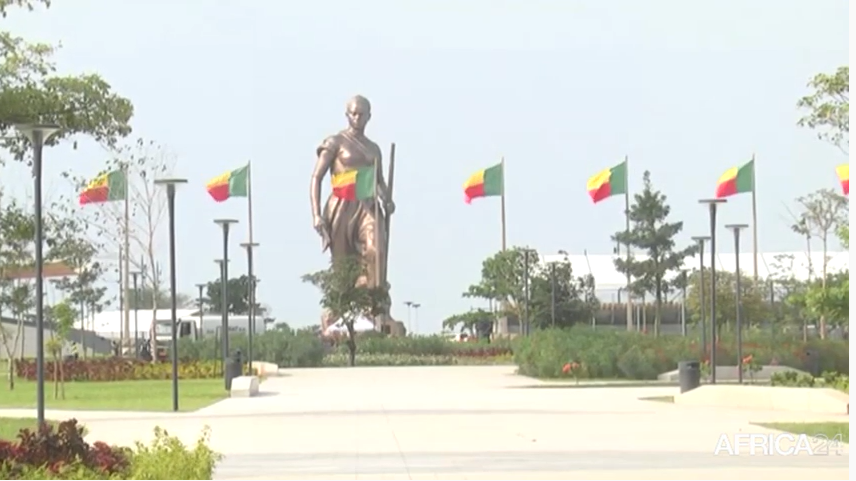 Grande statue debout tenant une lance entourée de dix drapeaux verts, jaunes et rouges le long d’une large allée plantée d’arbres et lampadaires