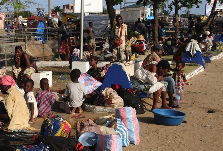 Groupe de personnes, adultes et enfants, assis et rassemblés avec leurs bagages dans un espace public extérieur