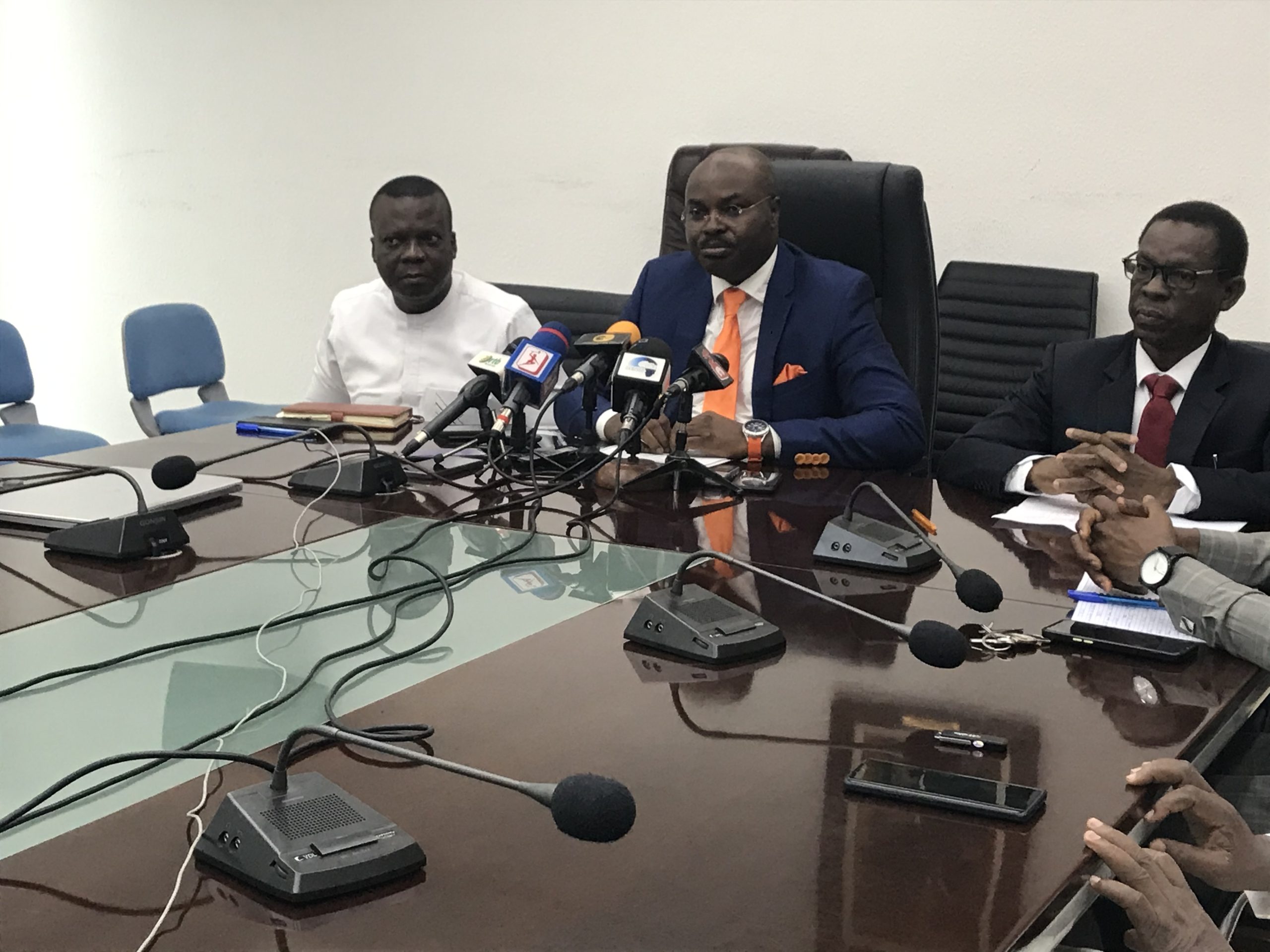 Trois hommes assis à une table de conférence avec plusieurs microphones et enregistreurs devant eux