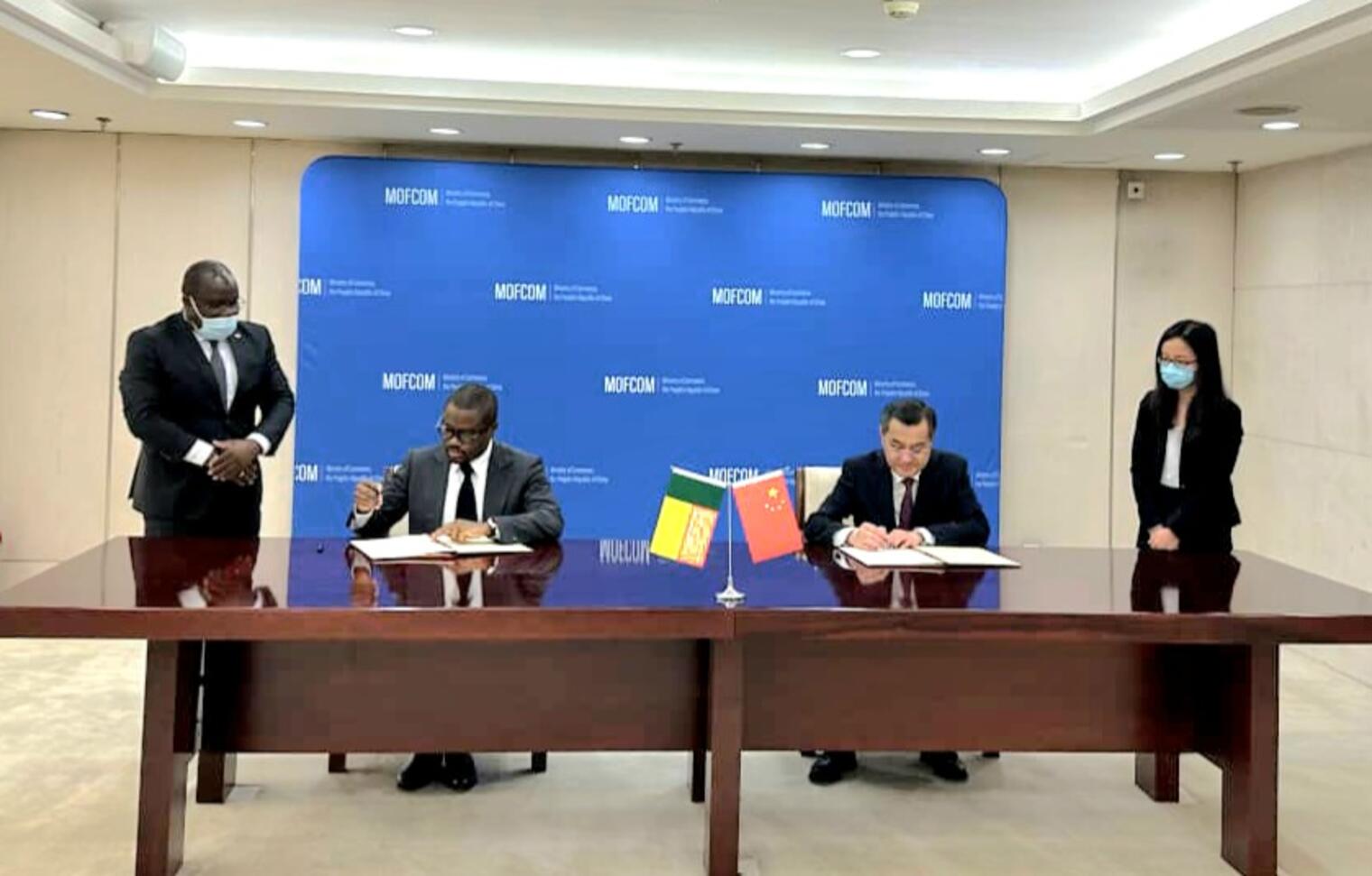 Deux hommes assis signent des documents derrière un bureau avec les drapeaux d’un pays africain et de la Chine au centre, deux personnes debout à côté.