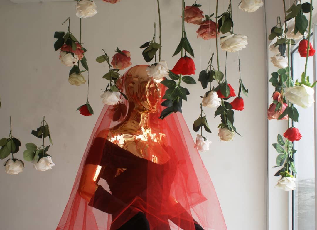 Buste doré drapé de tulle rouge sous des bouquets de roses suspendus aux tiges vertes sur fond clair