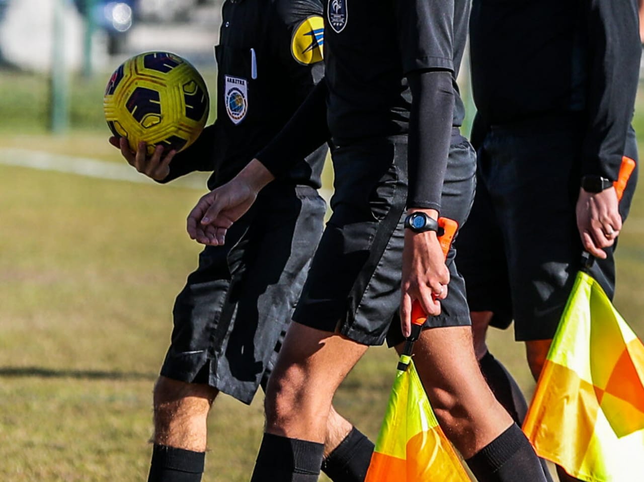 Trois arbitres en tenue noire marchent sur le terrain, un tenant un ballon jaune, deux portant des drapeaux de touche.