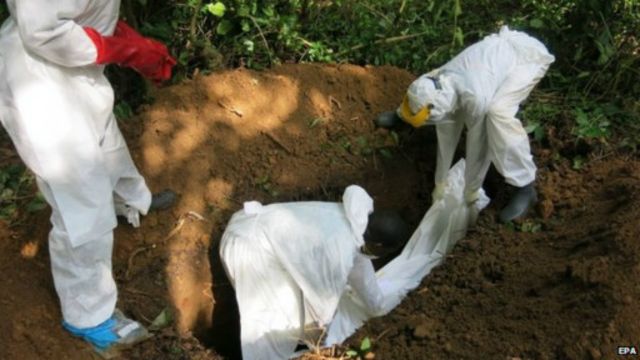 Trois personnes en combinaison de protection enterrent un corps dans une fosse creusée en pleine nature