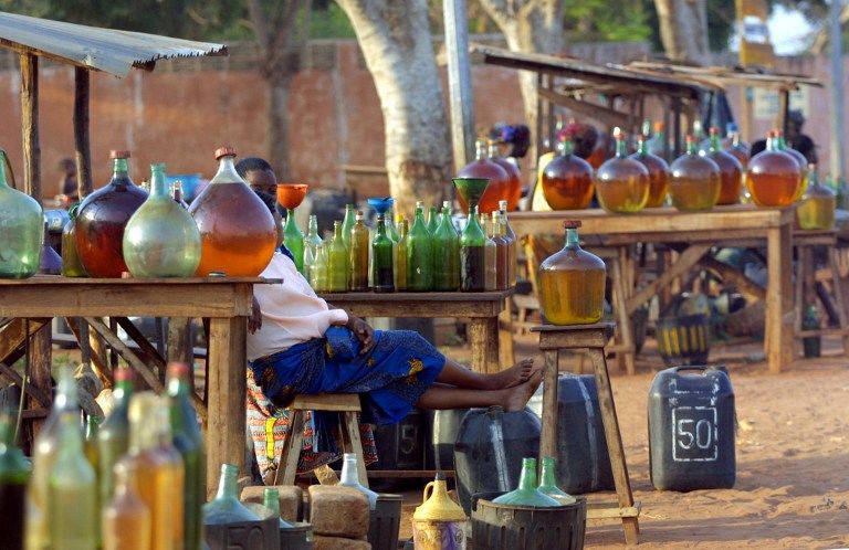 Vendeuse assise derrière un étal rempli de bouteilles et bidons colorés disposés sur des tables en plein air dans un marché rural
