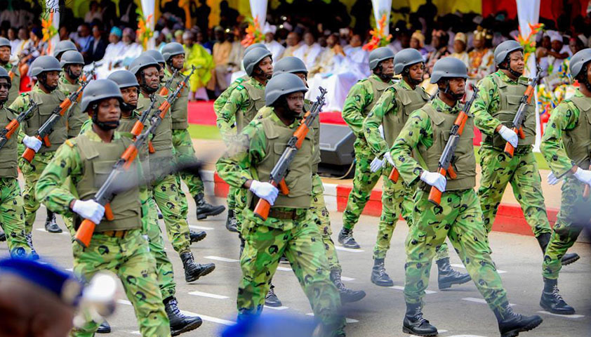 Des soldats en tenue camouflage verte et casques défilent dans une rue devant des spectateurs assis sous un chapiteau coloré
