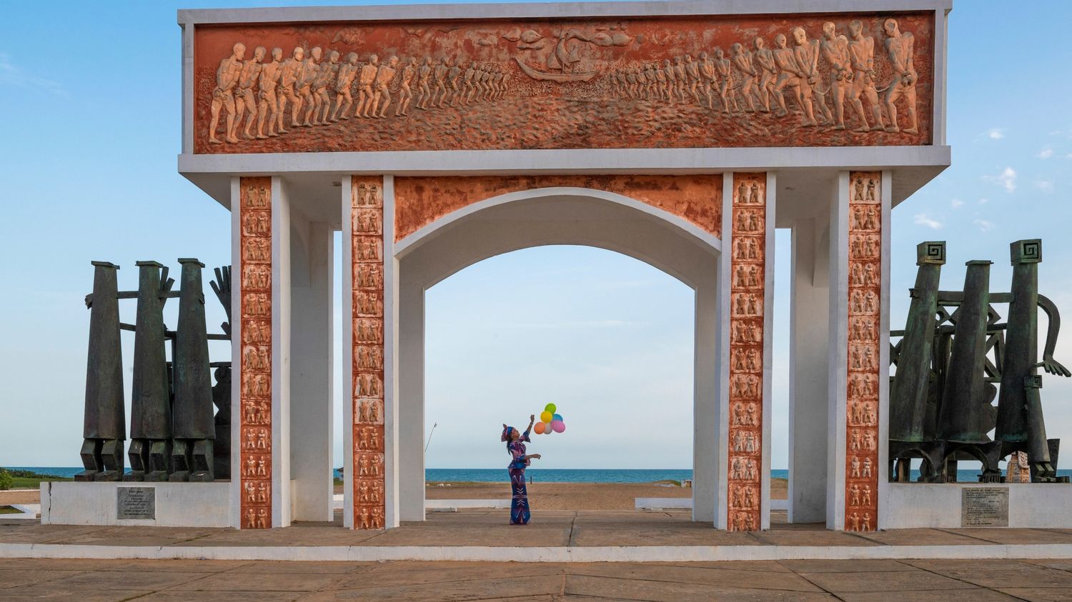 Monument avec sculptures en bas-relief encadrant une personne tenant des ballons colorés face à la mer