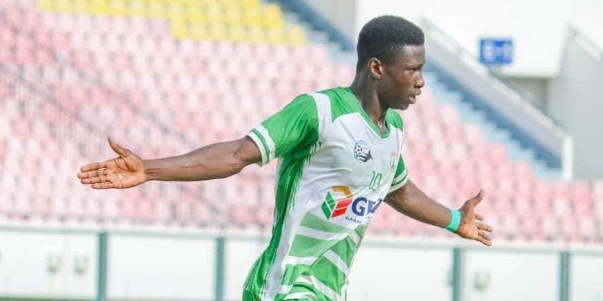 Un joueur de football en maillot vert et blanc étend les bras sur un terrain avec des tribunes vides en arrière-plan