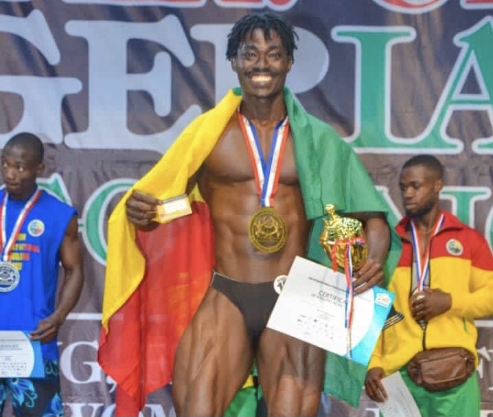 Un athlète musculaire souriant drapé dans un drapeau tient un trophée et un certificat sur scène devant deux autres concurrents médaille au cou