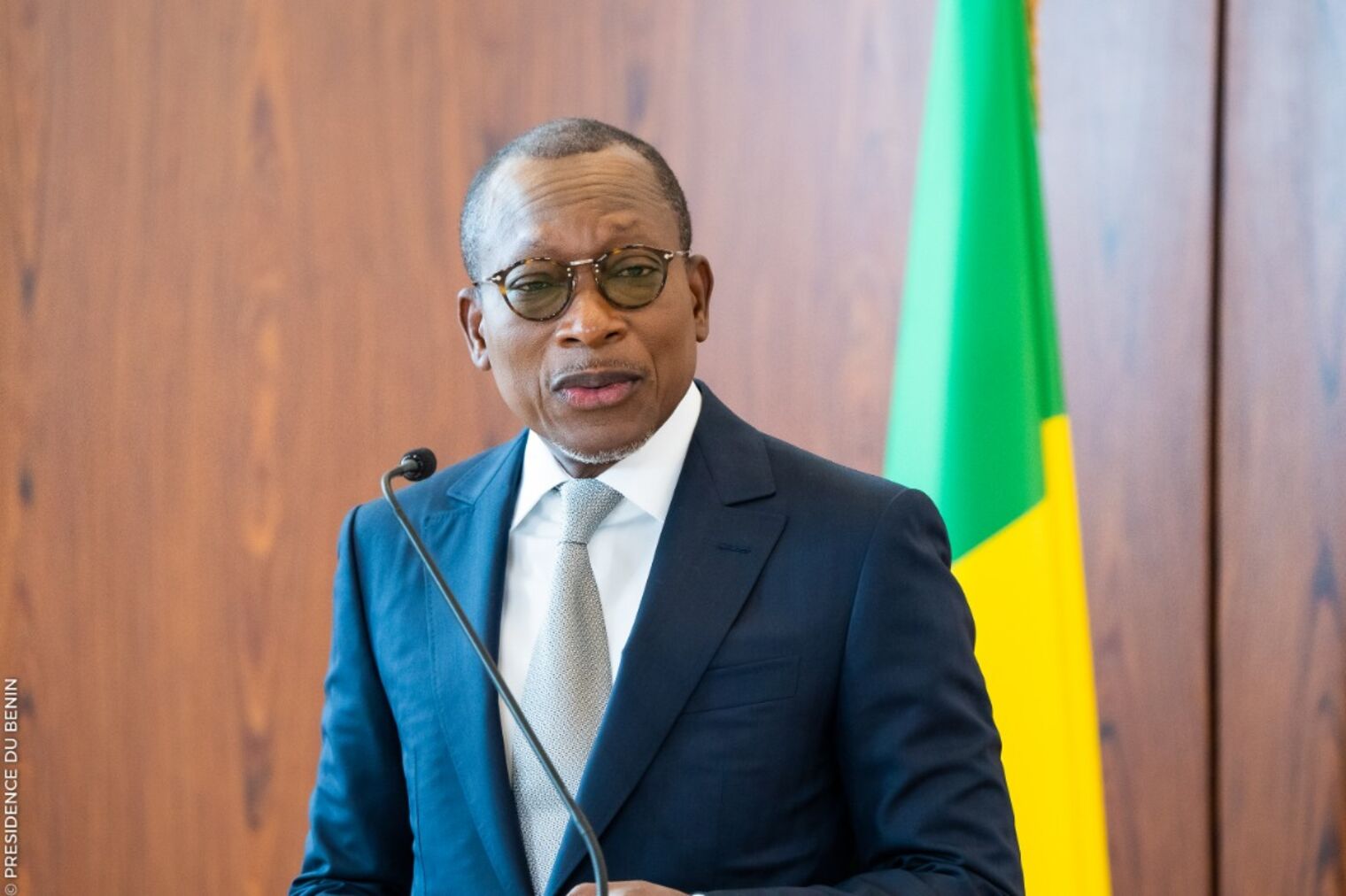 Un homme en costume sombre et cravate parle derrière un micro avec un drapeau jaune et vert en arrière-plan