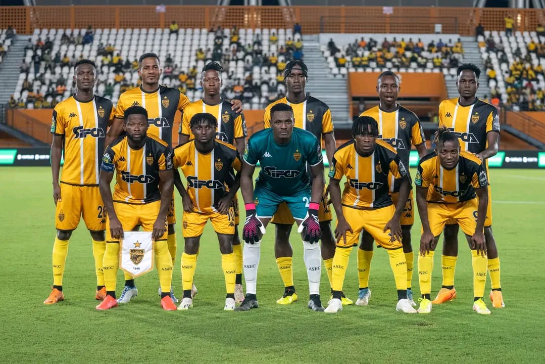 Onze joueurs de football en tenue jaune et noire posent sur un terrain de stade devant des tribunes partiellement remplies