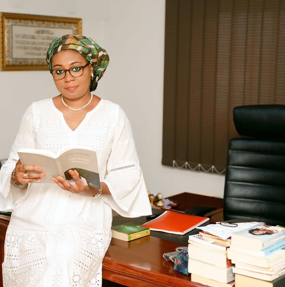 Femme en tenue traditionnelle blanche et foulard vert lisant un livre dans un bureau avec des piles de livres sur un bureau en bois