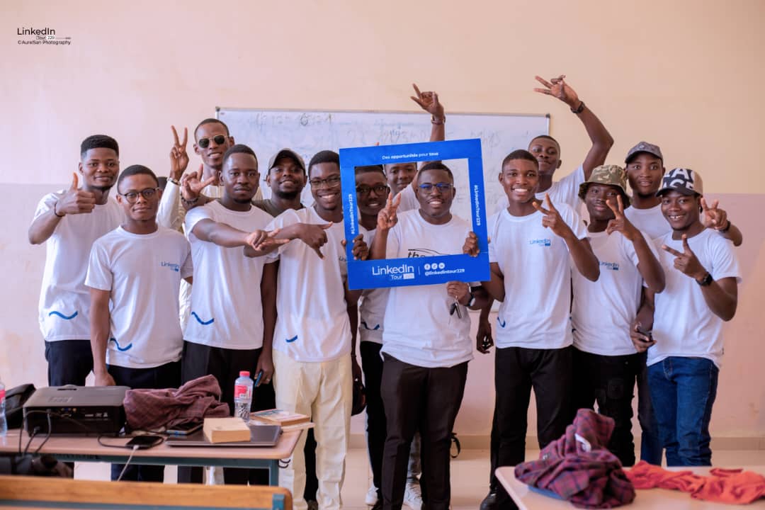 Un groupe de jeunes hommes souriants portent des t-shirts blancs et font des gestes de victoire en posant avec un cadre LinkedIn bleu dans une salle de classe