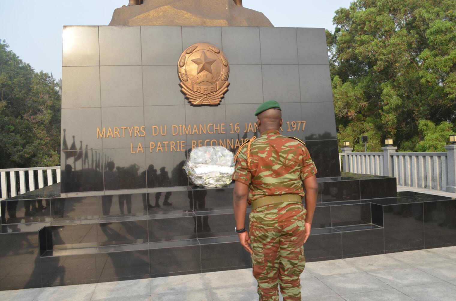 Un militaire en tenue camouflage et béret vert face à un monument commémoratif orné d’une gerbe de fleurs blanches