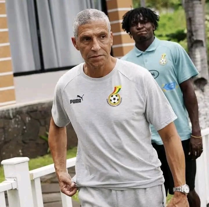 Un homme en tenue de sport grise avec un logo de football marche devant un autre homme en polo bleu clair dans un environnement extérieur.