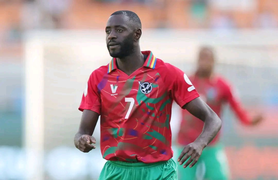 Joueur de football en maillot rouge et short vert sur un terrain avec un autre joueur flou en arrière-plan