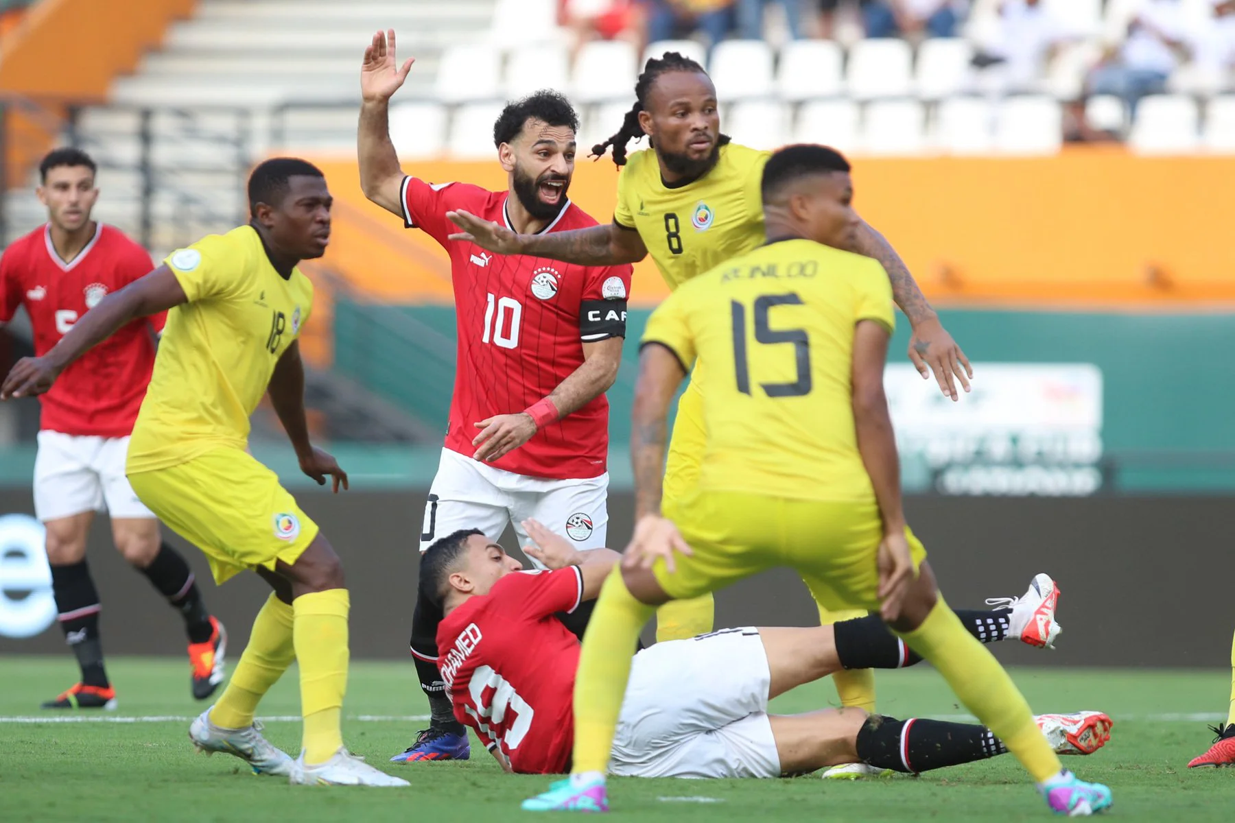 Joueur en rouge levant le bras entouré de quatre joueurs en jaune dont un au sol lors d'un match sur pelouse verte