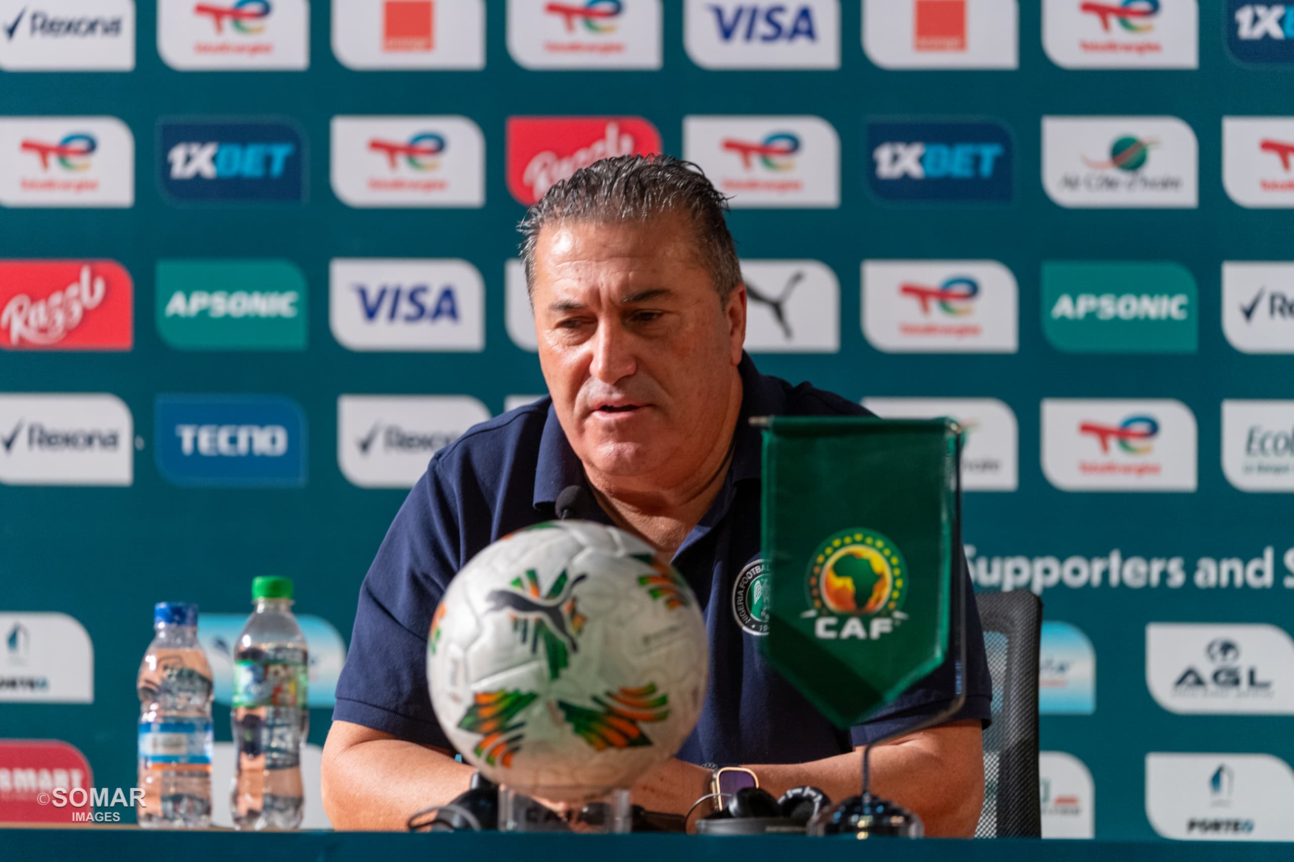 Un homme assis à une table de conférence avec un ballon de football et une petite bannière à son effigie devant lui