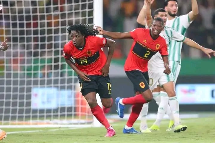 Deux joueurs en maillot rouge et noir courent devant un gardien et un défenseur en blanc et vert près d'un but de football