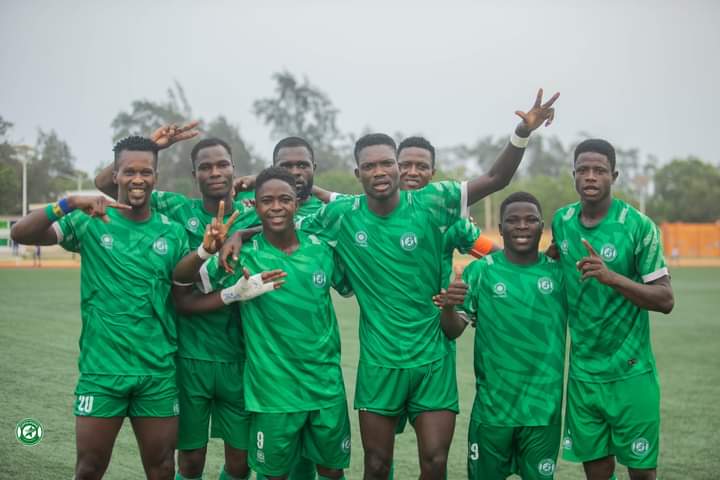 Sept joueurs vêtus de maillots verts posent bras dessus bras dessous en souriant sur un terrain de football.