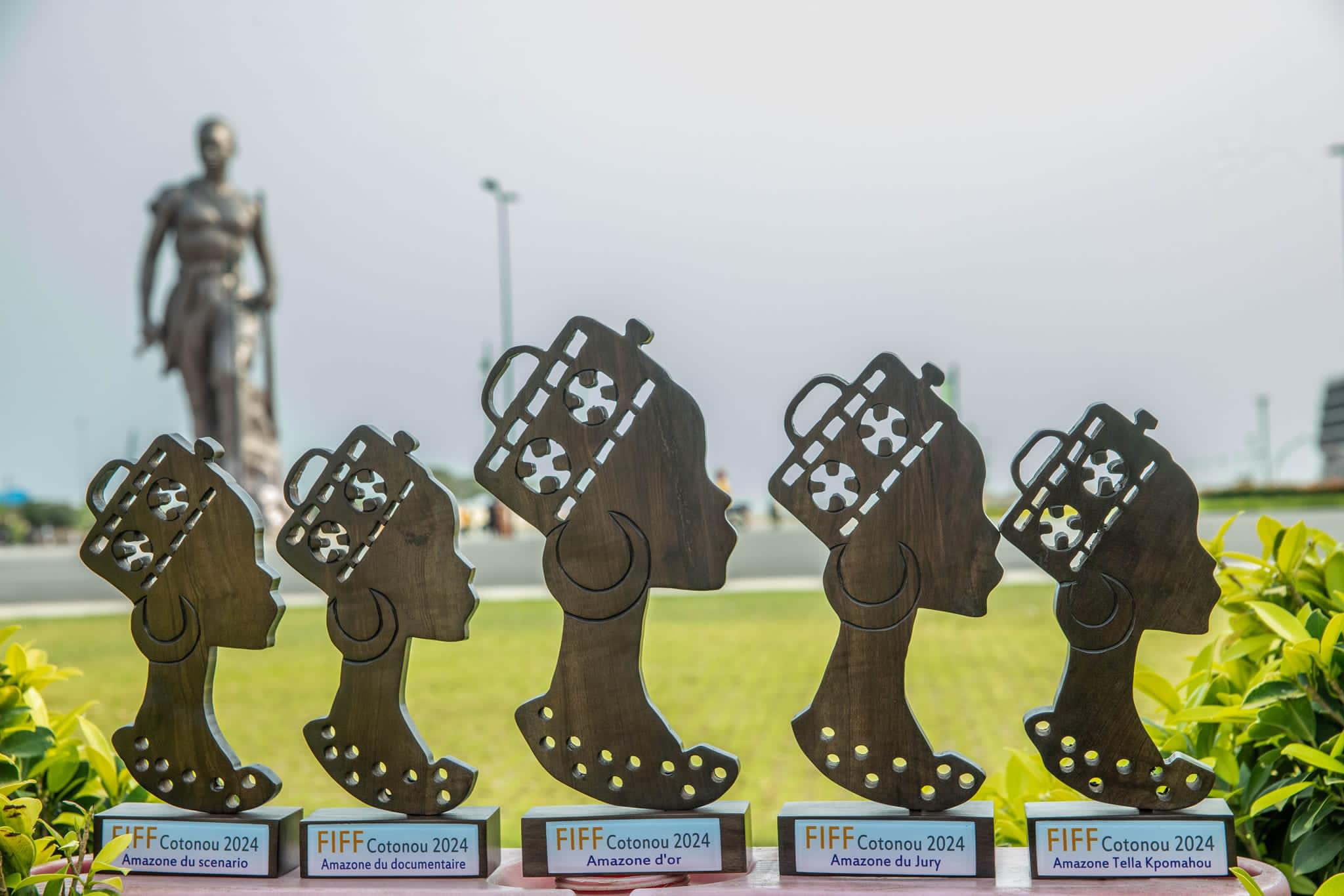 Cinq trophées en forme de silhouette féminine ornée d'une coiffe, posés sur une surface avec un jardin et une statue floue en arrière-plan