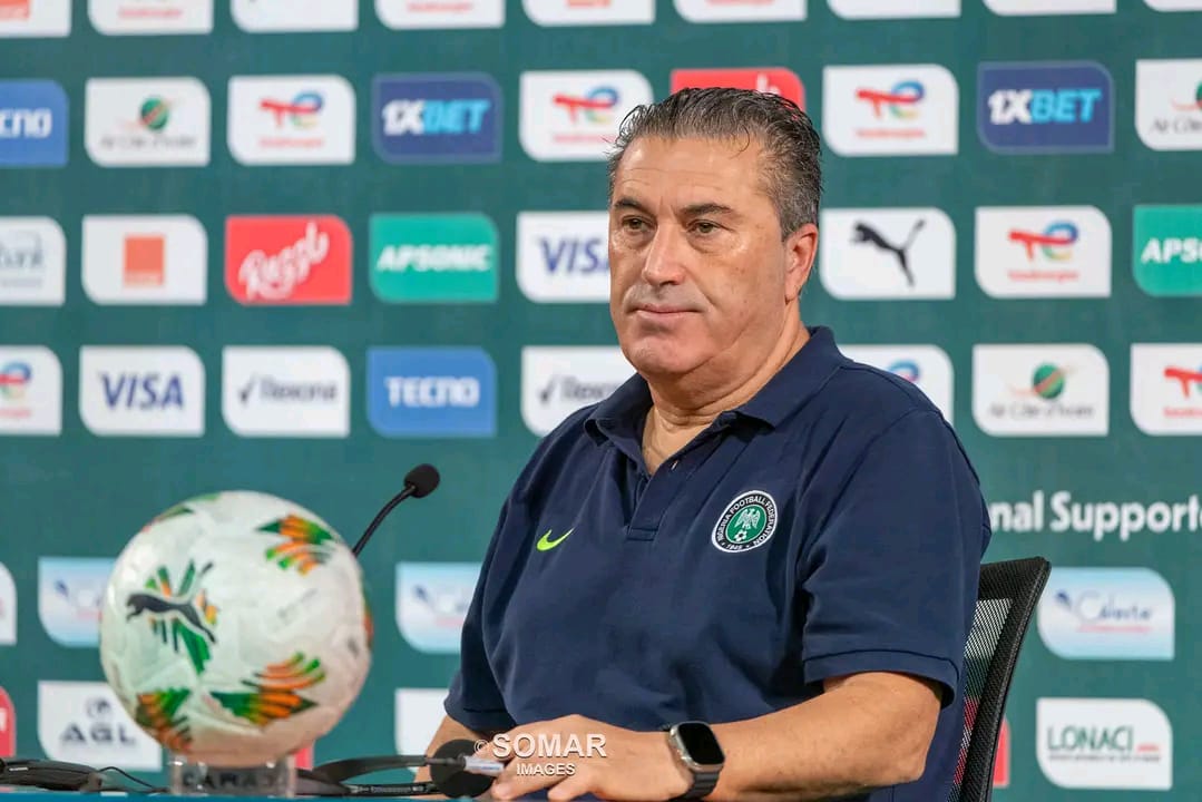 Homme en polo bleu marine assis devant un micro et un ballon de football lors d une conférence de presse