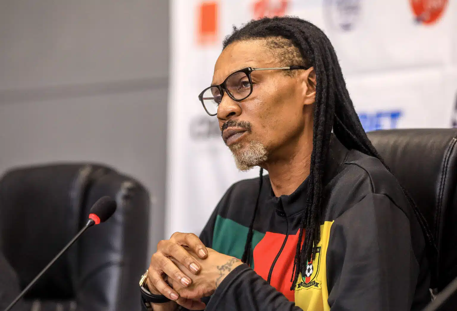 Homme aux dreadlocks et lunettes assis les mains jointes devant un micro en conférence de presse avec un blason football sur son sweat