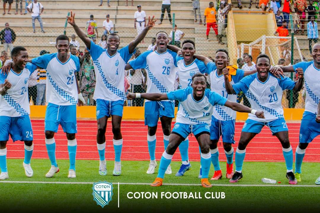 Une équipe de football en maillots blancs et bleus célèbre ensemble sur un terrain gazonné devant des gradins partiellement remplis