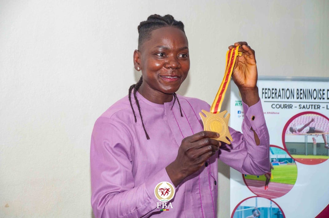 Une femme souriante en chemise mauve tient une médaille dorée avec un ruban jaune et rouge devant un poster sportif