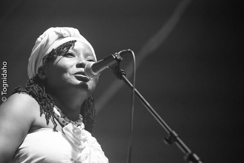 Femme en tenue blanche coiffée d'un turban chantant devant un micro sur scène avec un éclairage tamisé