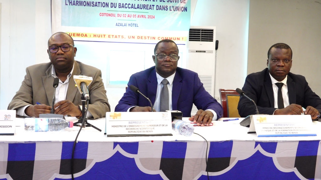Trois hommes en costume assis derrière une table avec des microphones et des panneaux nominaux lors d'une réunion officielle