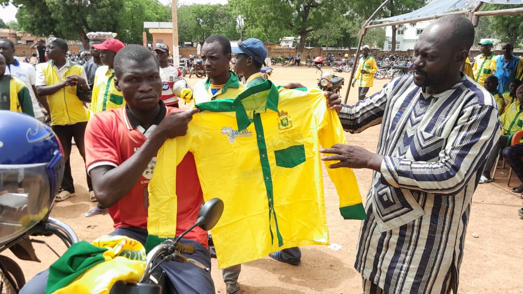 Un homme tend une chemise jaune à rayures vertes à un autre homme en polo rouge dans un rassemblement en plein air