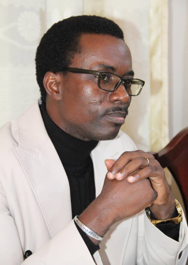 Homme portant des lunettes et un pardessus clair, les mains jointes, regard pensif vers la droite
