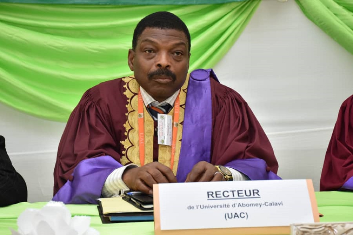 Un homme en toge universitaire violette et or est assis derrière une table avec un panneau « RECTEUR » et un badge visible au cou