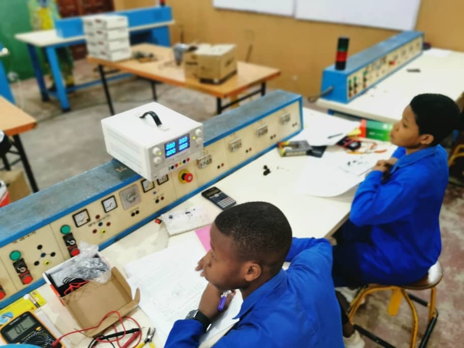 Deux étudiants en blouse bleue sont assis à une table d’atelier avec équipements électroniques et documents techniques
