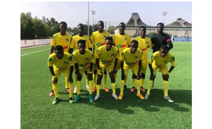 Onze jeunes joueurs de football en tenues jaunes posent sur un terrain herbeux devant des bâtiments et des arbres