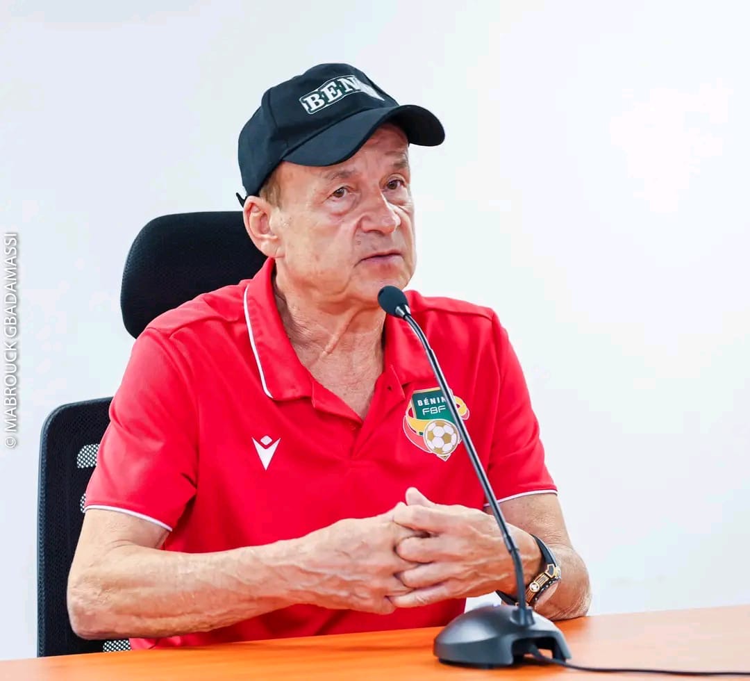 Homme portant une casquette et un polo rouge avec un blason, parlant devant un micro posé sur une table en bois claire