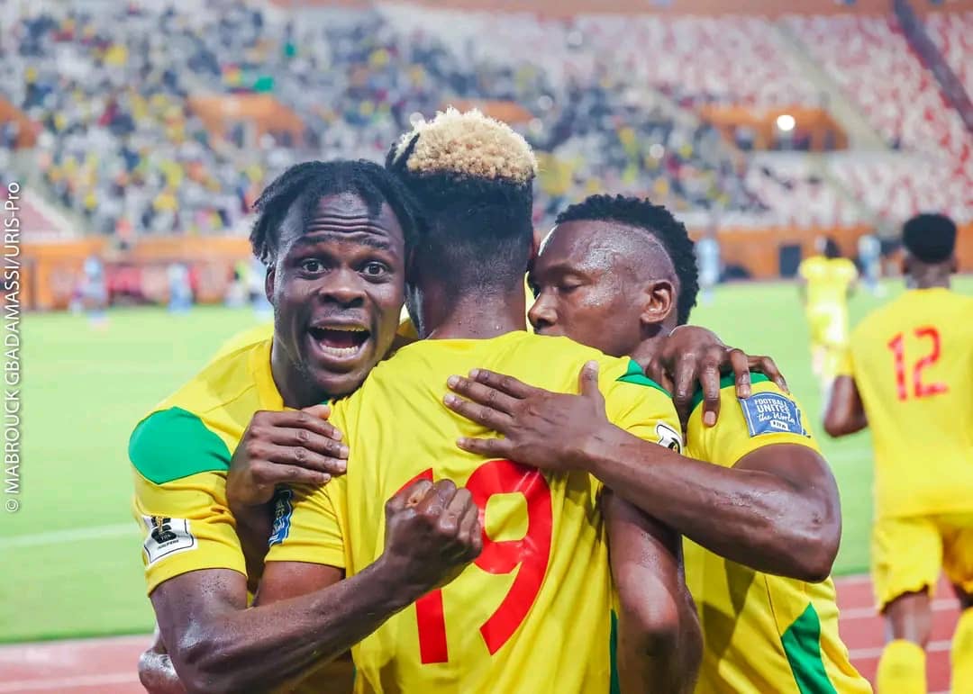 Trois joueurs en maillots jaunes s’embrassent et célèbrent sur un terrain de football devant un public en tribunes