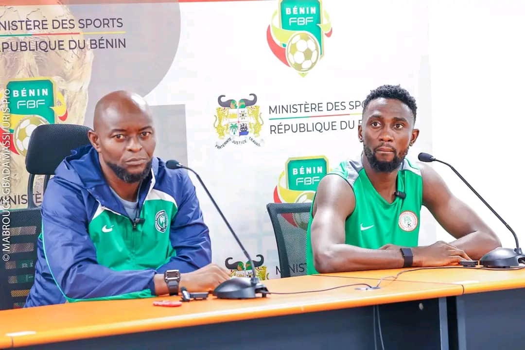 Deux hommes assis à une table de conférence avec microphones et logos sportifs en arrière-plan