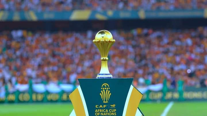 Trophée doré de la Coupe d'Afrique des Nations sur un socle vert et jaune avec un stade flou en arrière-plan