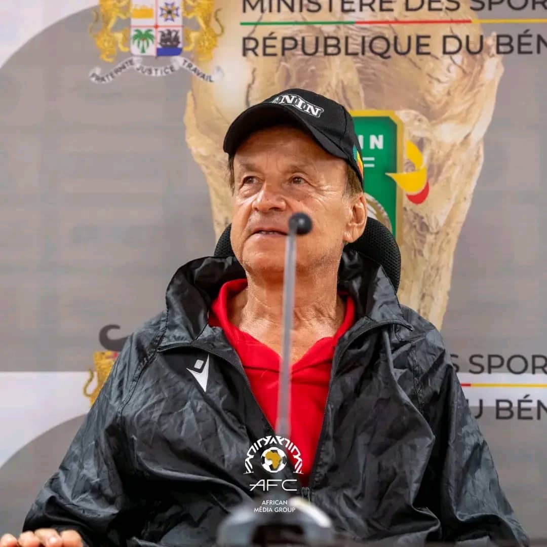 Homme assis en veste noire et casquette, micro devant lui, devant un panneau officiel du ministère des sports