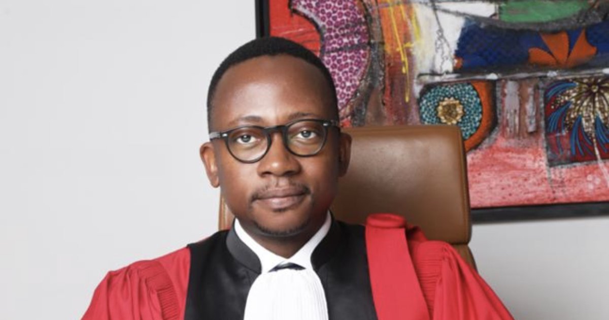 Homme en tenue d’avocat noire et rouge assis devant un mur décoré d’un tableau coloré