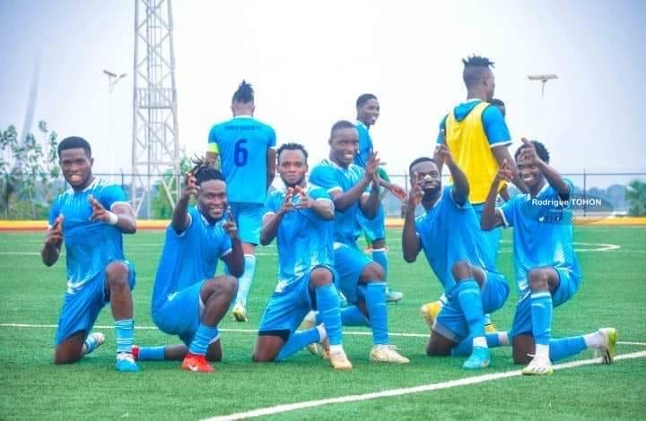 Joueurs en tenue bleue célébrant à genoux et debout sur un terrain de football gazonné sous un ciel clair