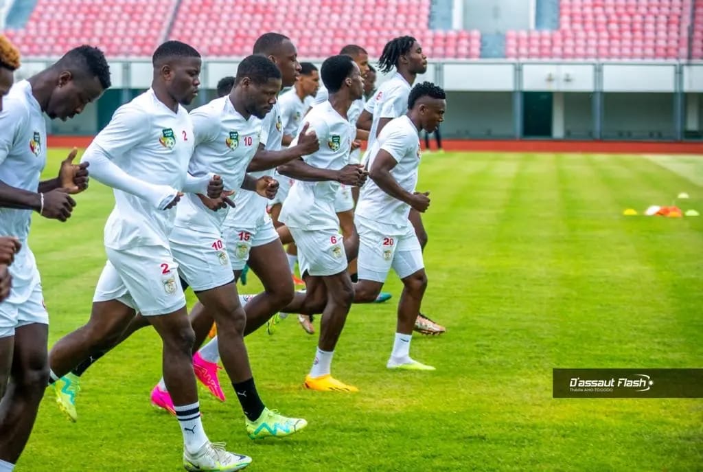 Des joueurs de football en tenue blanche effectuent des exercices de course sur un terrain vert dans un stade aux gradins rouges.