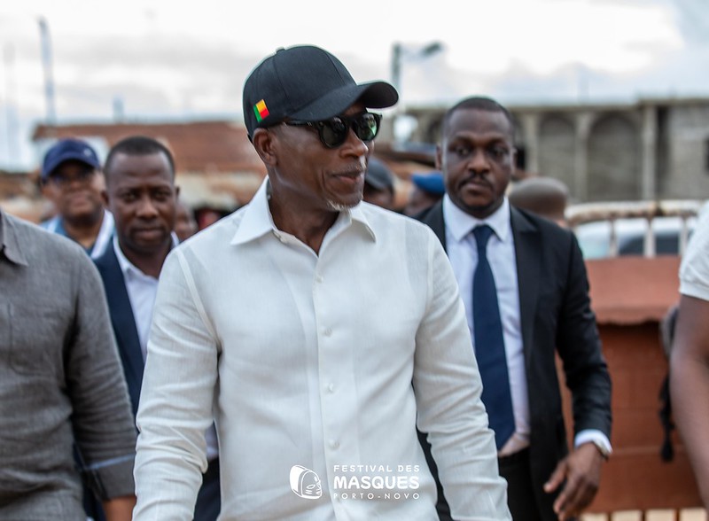 Un homme en chemise blanche, casquette noire et lunettes de soleil marche entouré de plusieurs hommes en arrière-plan