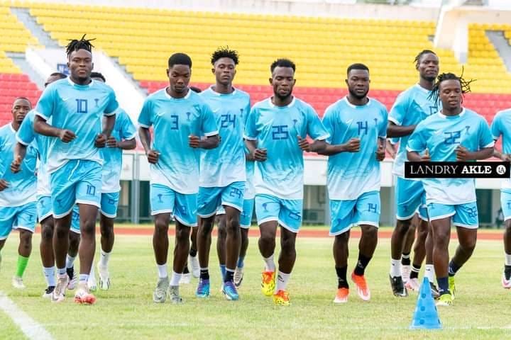 Un groupe de joueurs de football en tenue bleu clair s'entraîne en courant sur un terrain devant des gradins colorés