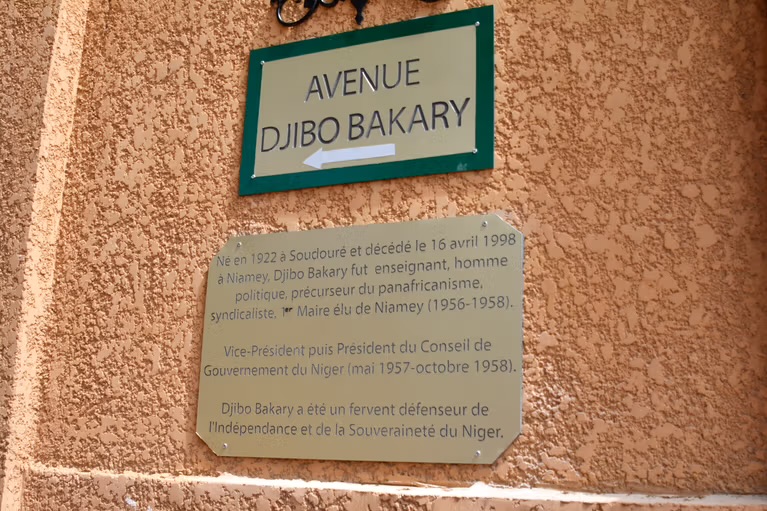 Plaque de rue verte indiquant Avenue Djibo Bakary avec une plaque explicative sur un mur ocre