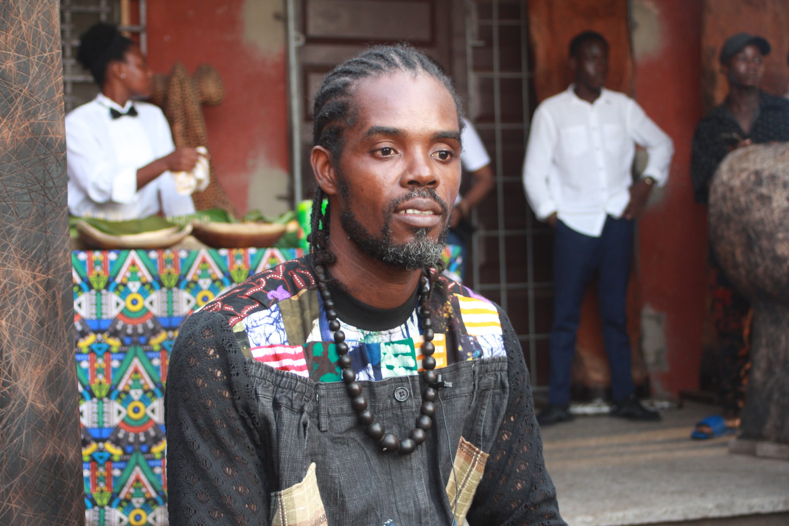 Homme avec des dreadlocks et un collier de perles assis devant un bâtiment entouré de plusieurs personnes en arrière-plan