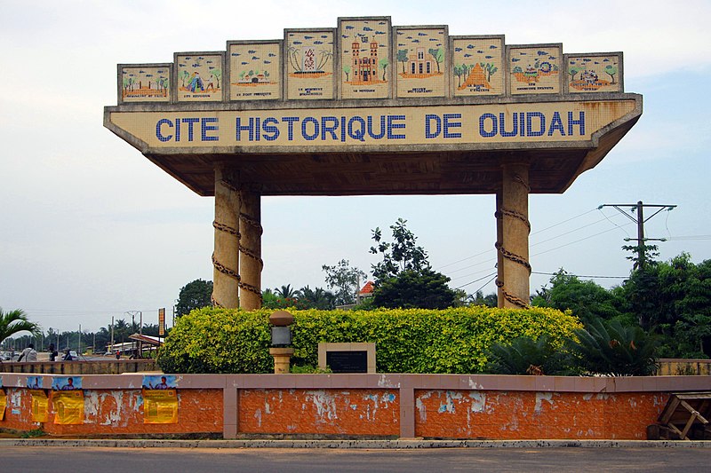 Panneau monumental en pierre indiquant une cité historique entourée de végétation et bordée par une route avec des affiches collées