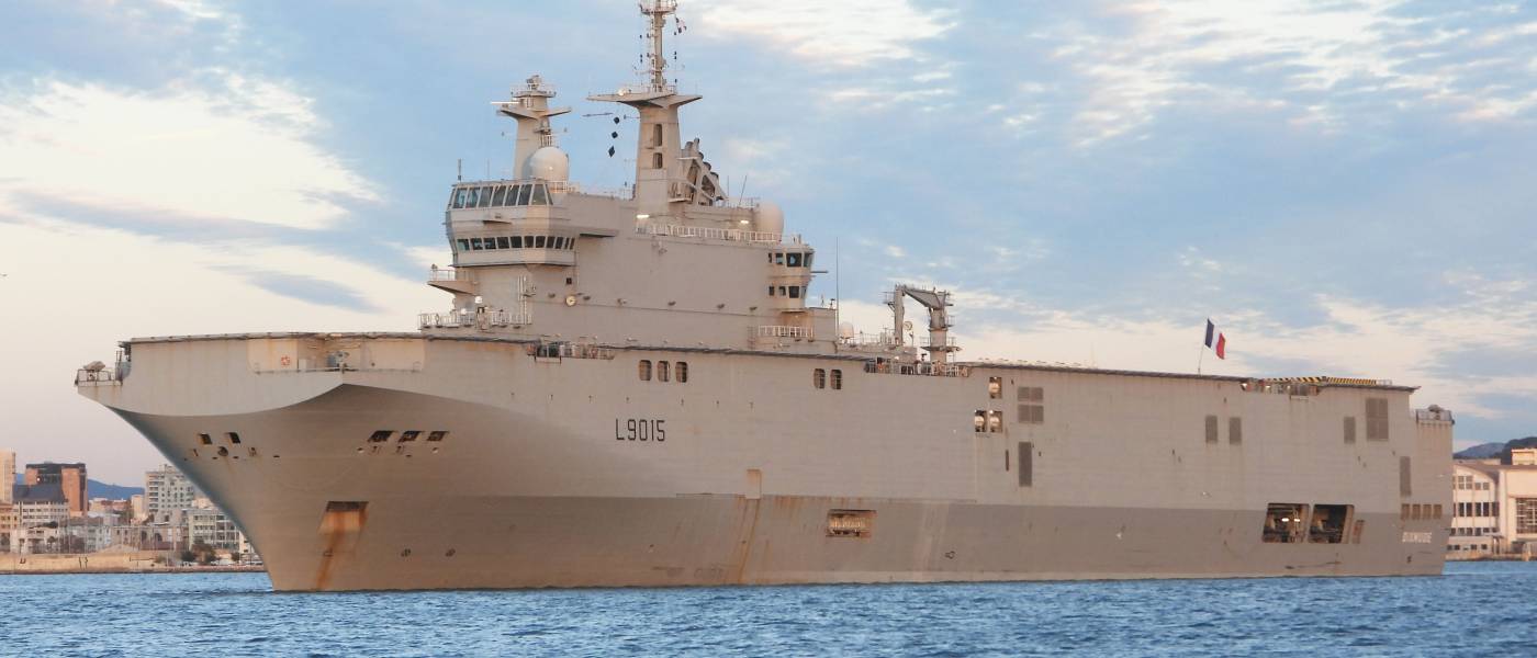 Navire militaire gris avec des inscriptions sur le côté amarré dans un port urbain sous ciel partiellement nuageux